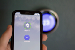 A hand holding up a smartphone in front of a smart thermostat